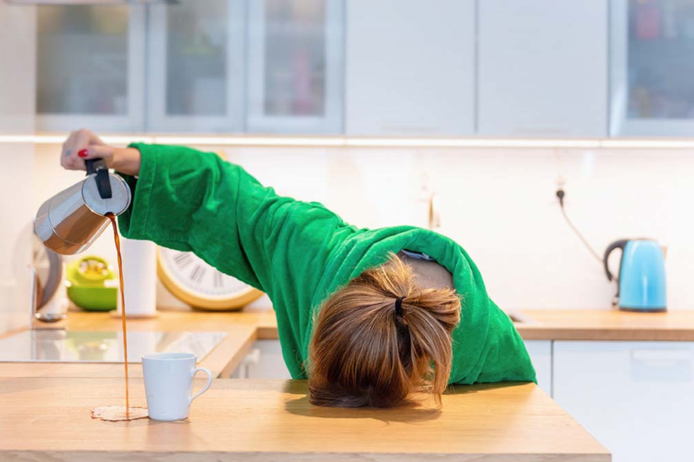 Een vrouw in een groen shirt die met haar hoofd op de tafel ligt, terwijl ze met een hand koffie naast een kopje schenkt.